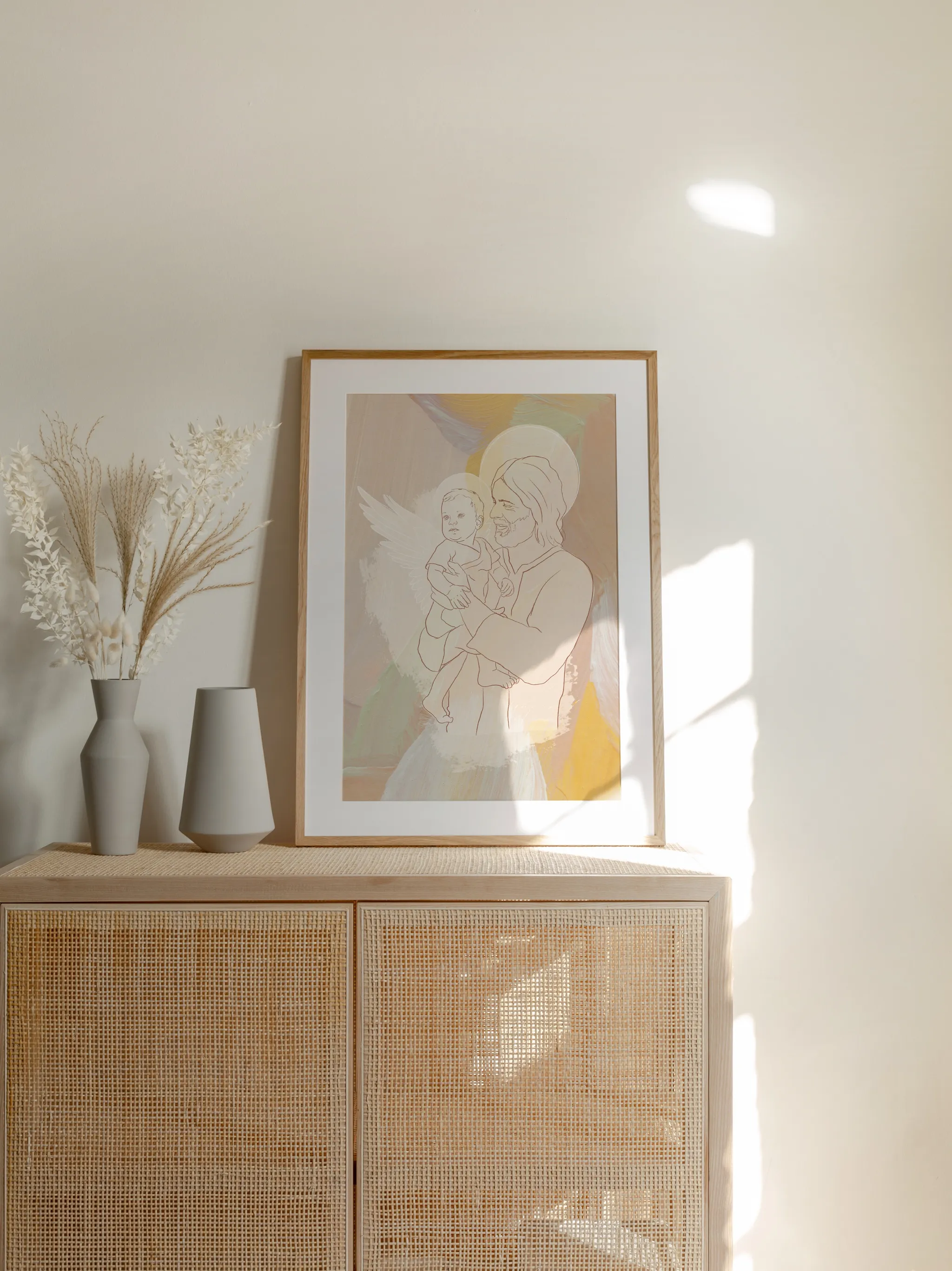 Angel Baby with Christ, framed in light wood with a white mat, standing on a rattan cabinet beside dried grasses and pale vases in sunlight.