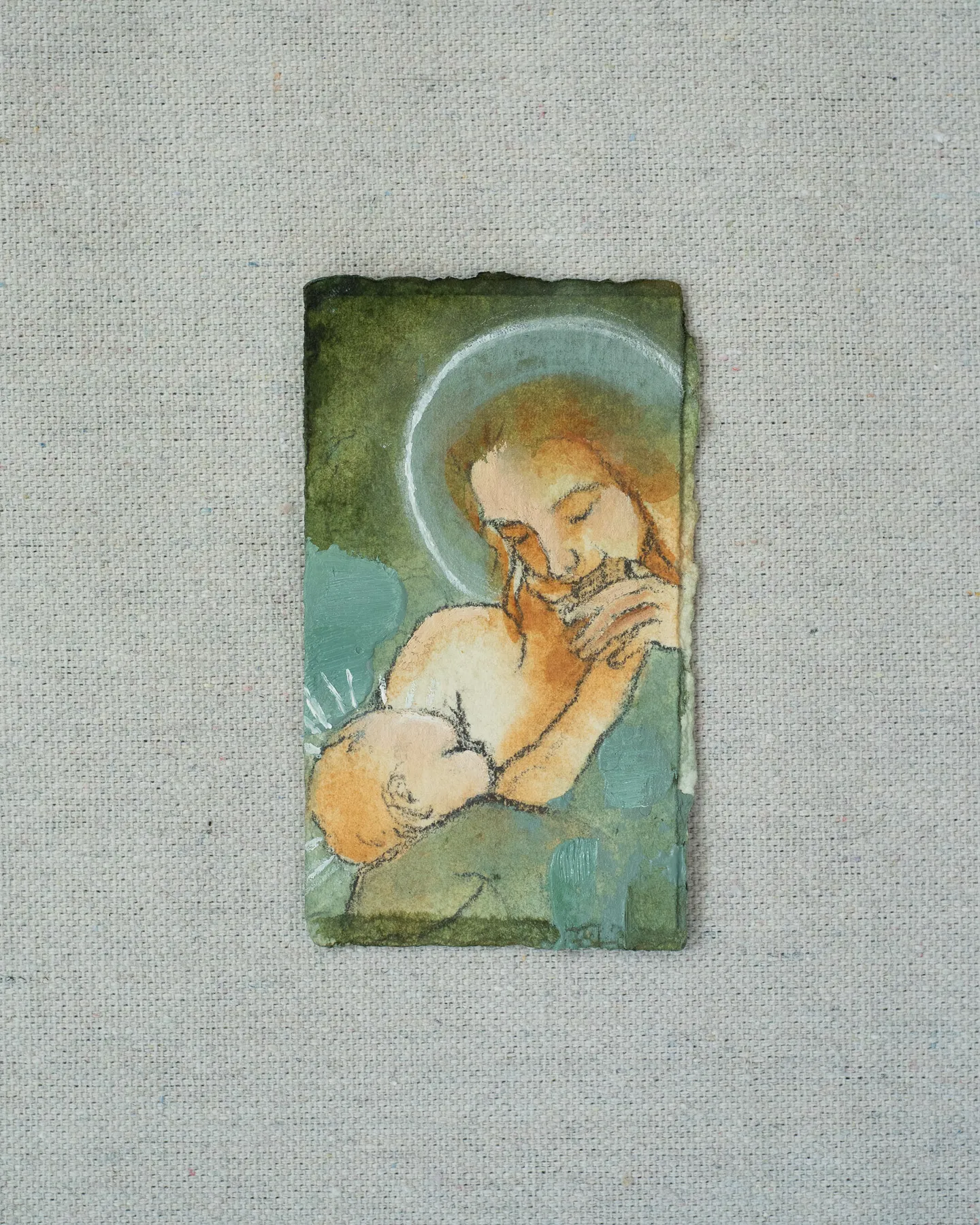 A small painting on torn-edged paper, laid on linen: a haloed mother with long auburn hair nursing her baby, in moss green and warm ochre.