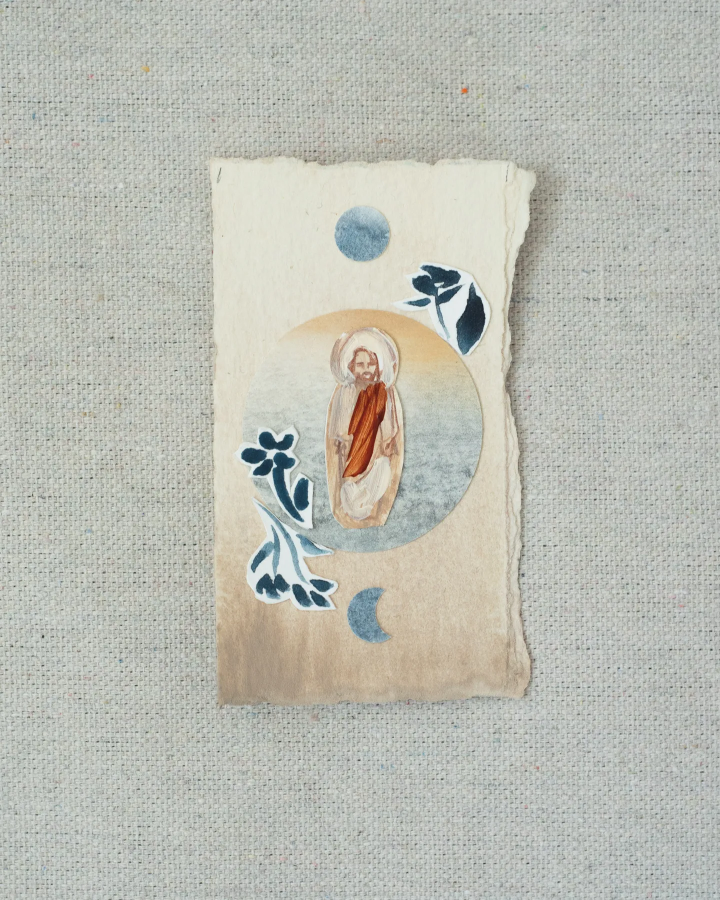 A small painting on torn-edged cream paper, laid on linen: a haloed figure of Christ in a red robe inside a pale blue circle, with a blue full moon above, a crescent below, and cut-paper indigo flowers.
