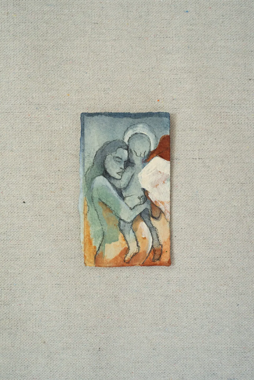 A small painting laid on linen, in slate blue and rust: a woman with long hair and a haloed figure holding each other close.