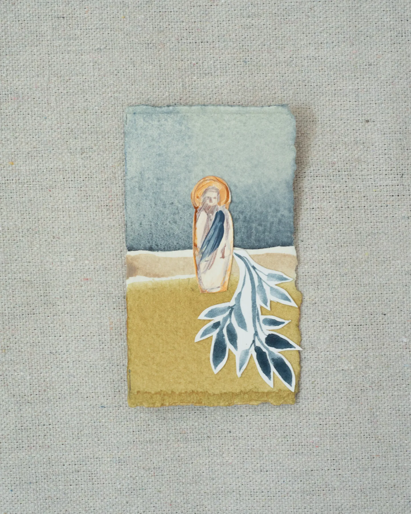 A small painting on torn-edged paper, laid on linen: a figure of Christ with a gold halo and a blue robe on a mustard field under a slate blue sky, with a cut-paper indigo branch at his feet.