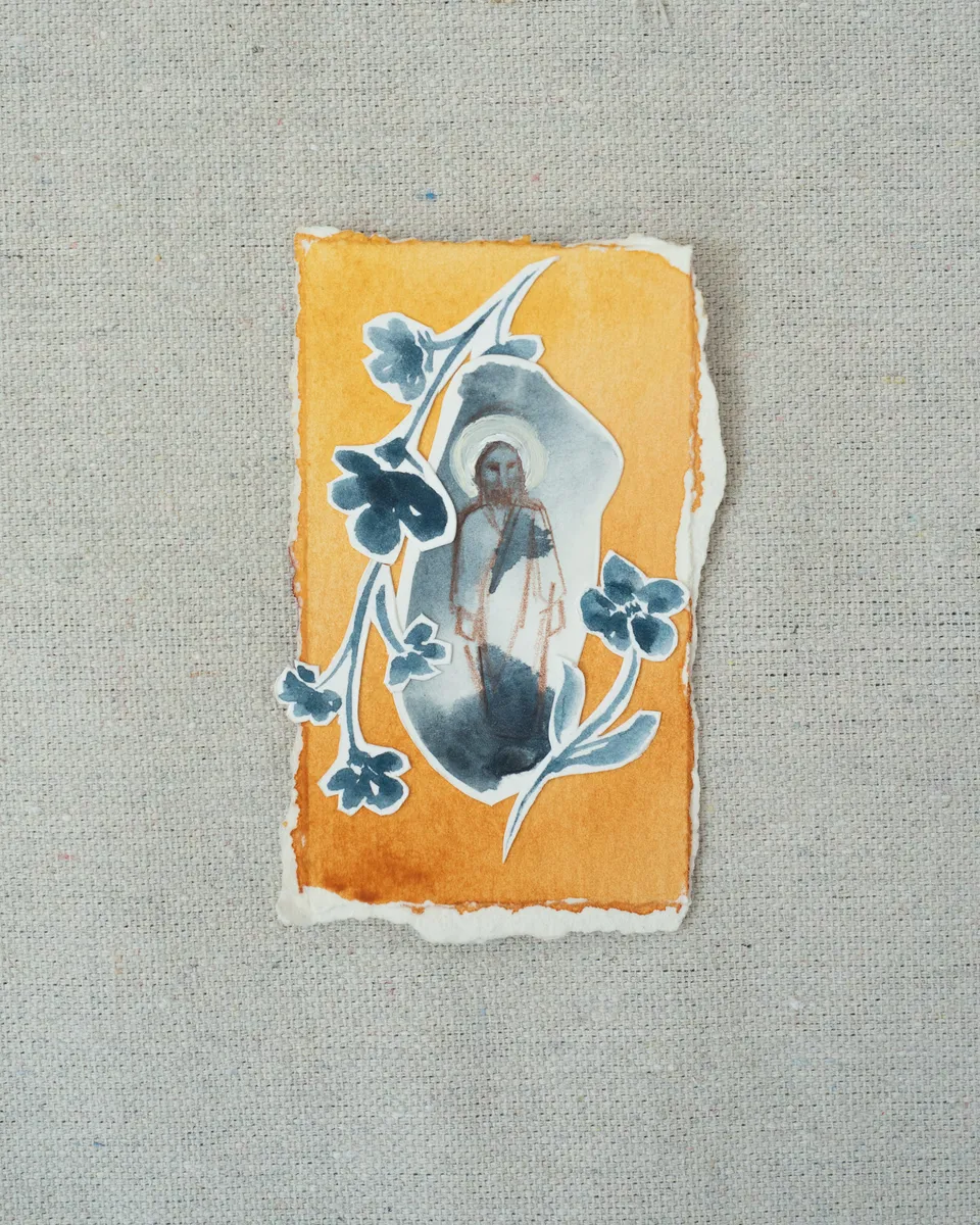 A small painting on torn-edged paper, laid on linen: a grey-blue figure of Christ with a halo on marigold orange, framed by a cut-paper vine of indigo flowers.
