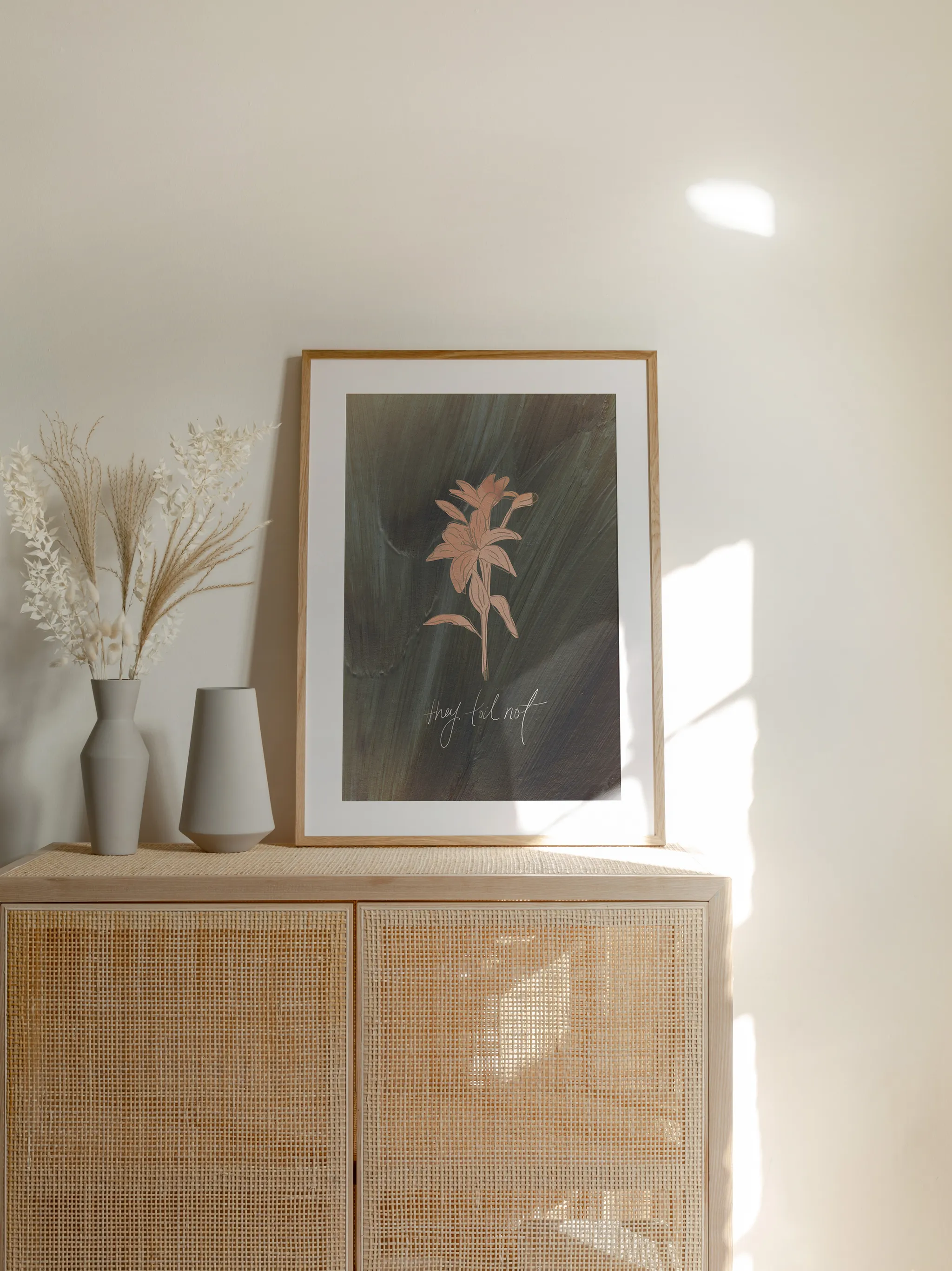 Consider the Lilies on Green, framed in light wood with a white mat, standing on a rattan cabinet beside dried grasses and pale vases in sunlight.
