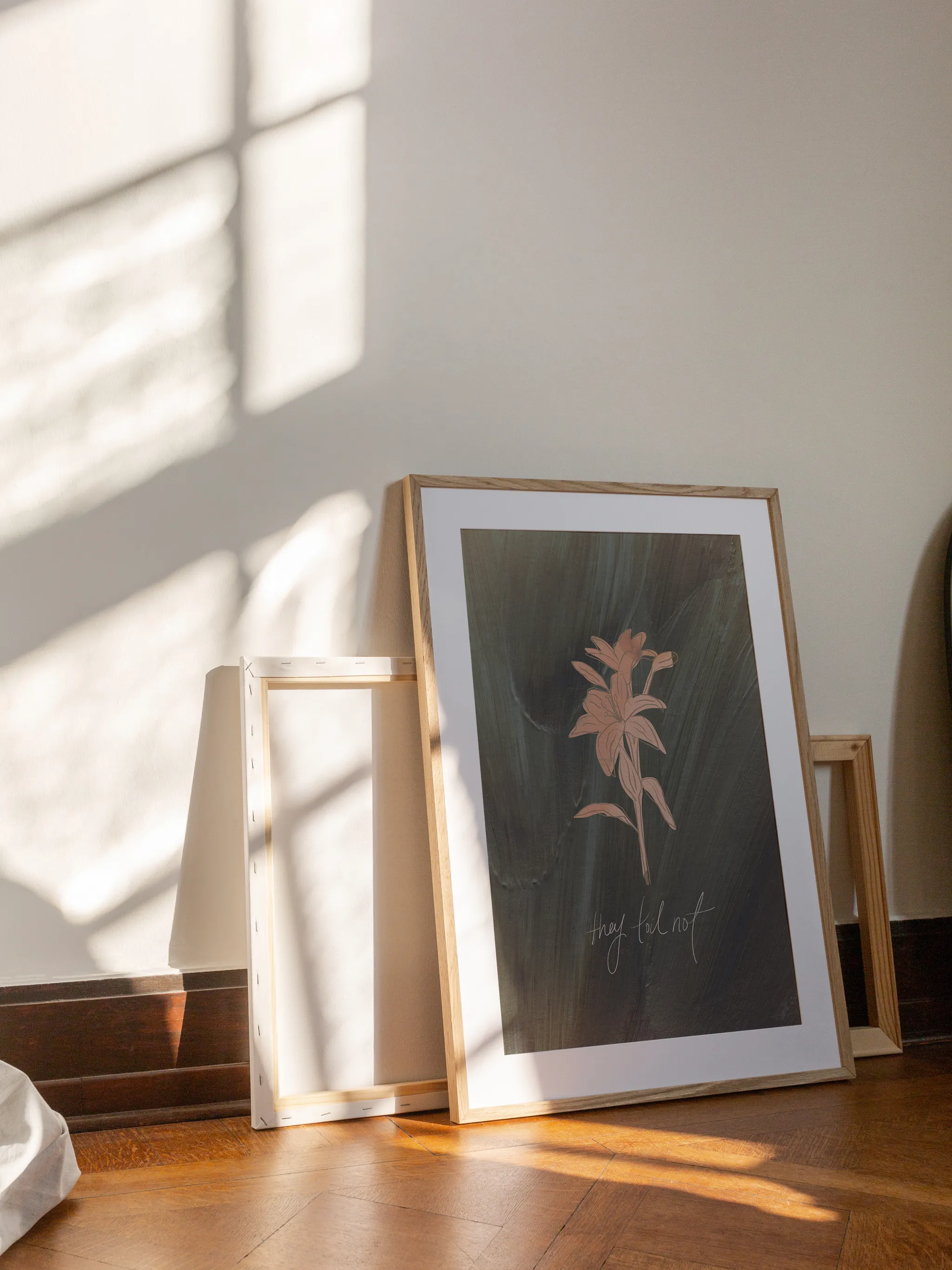 Consider the Lilies on Green, framed in light wood with a white mat, leaning against a wall on a wooden floor in slanting sunlight, with empty frames behind it.