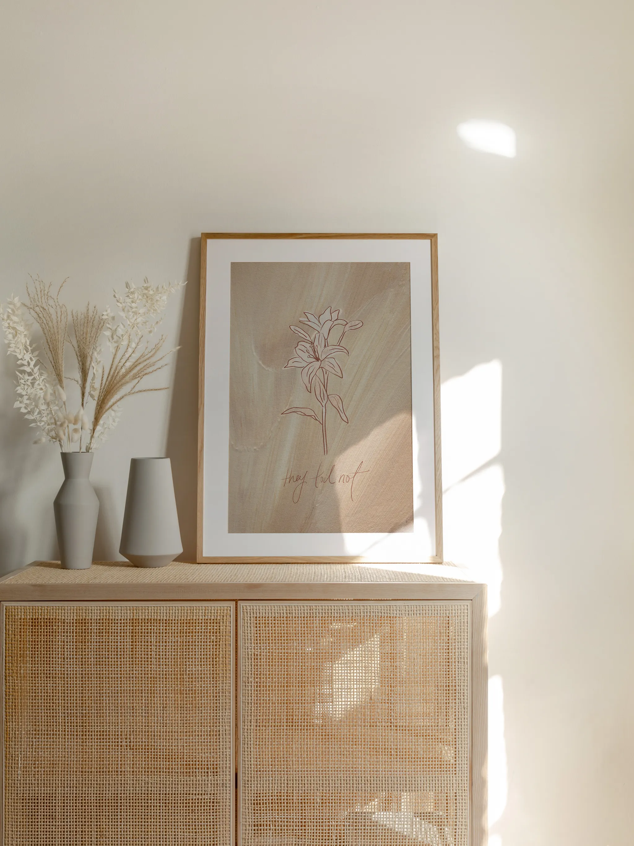 Consider the Lilies on Tan, framed in light wood with a white mat, standing on a rattan cabinet beside dried grasses and pale vases in sunlight.