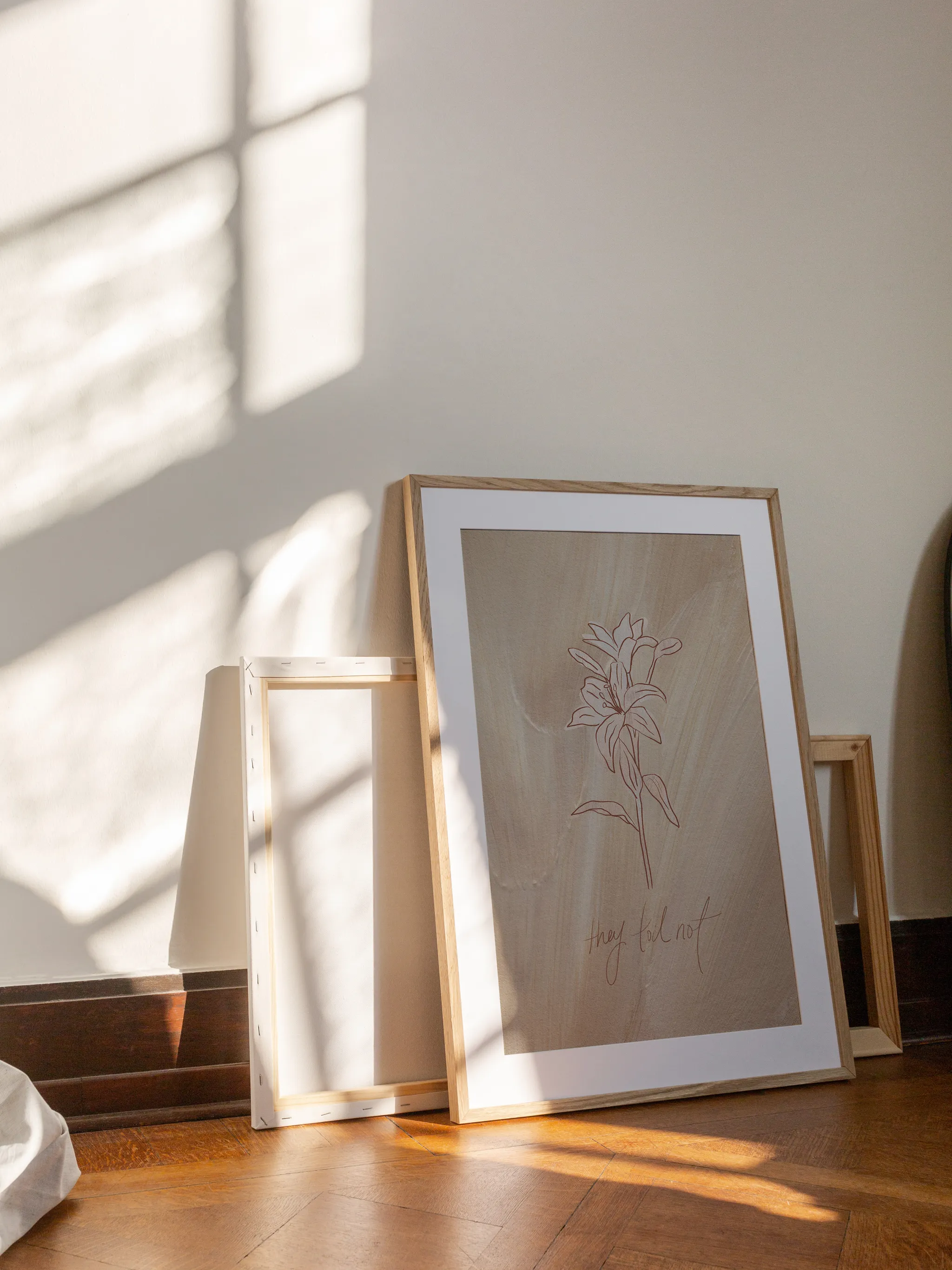Consider the Lilies on Tan, framed in light wood with a white mat, leaning against a wall on a wooden floor in slanting sunlight, with empty frames behind it.