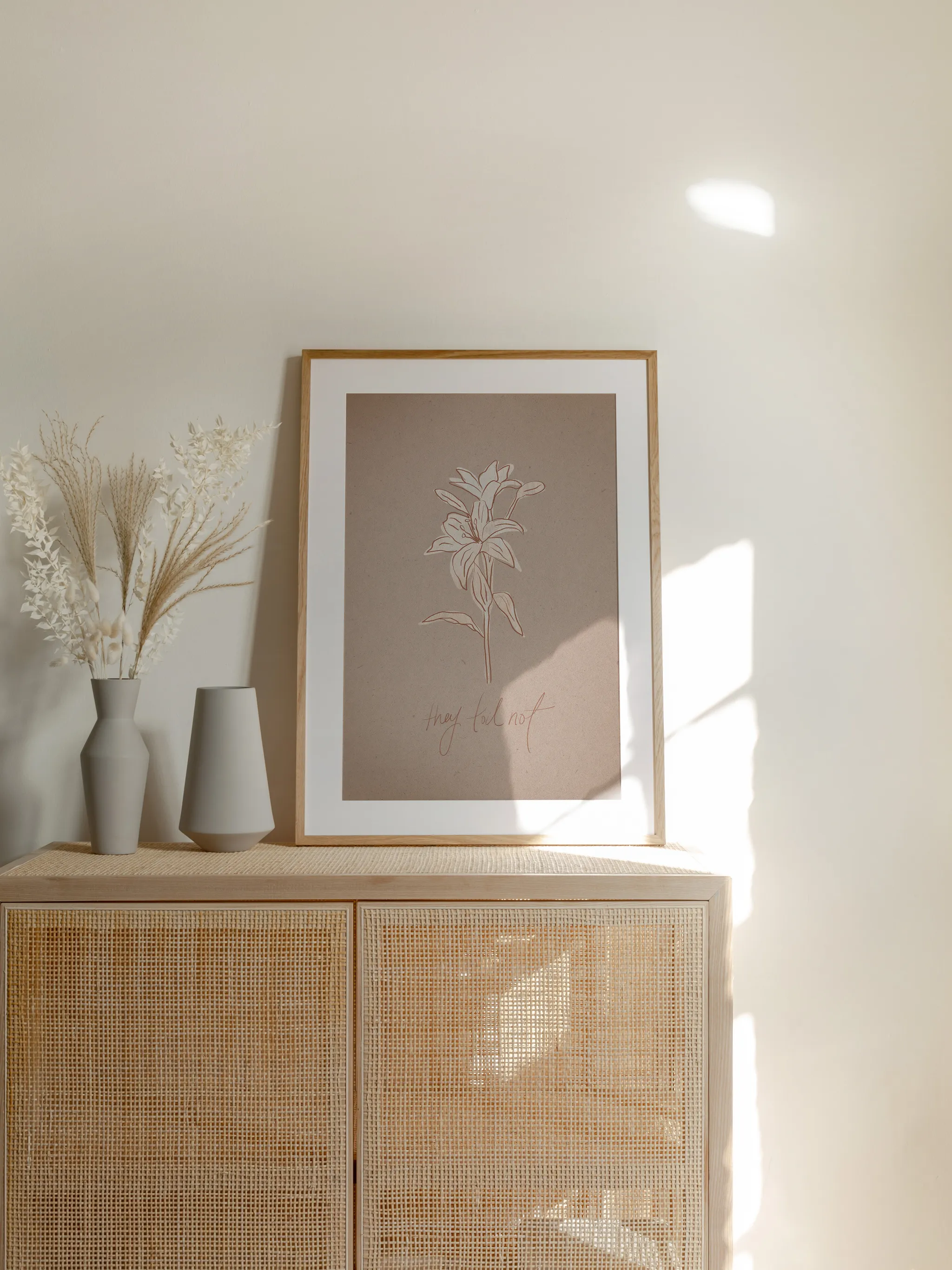 Consider the Lilies on Paper, framed in light wood with a white mat, standing on a rattan cabinet beside dried grasses and pale vases in sunlight.