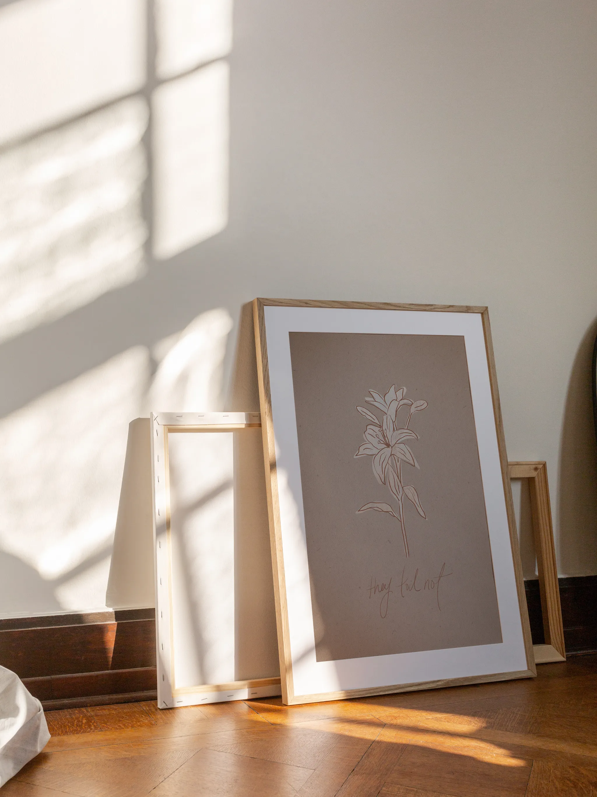 Consider the Lilies on Paper, framed in light wood with a white mat, leaning against a wall on a wooden floor in slanting sunlight, with empty frames behind it.