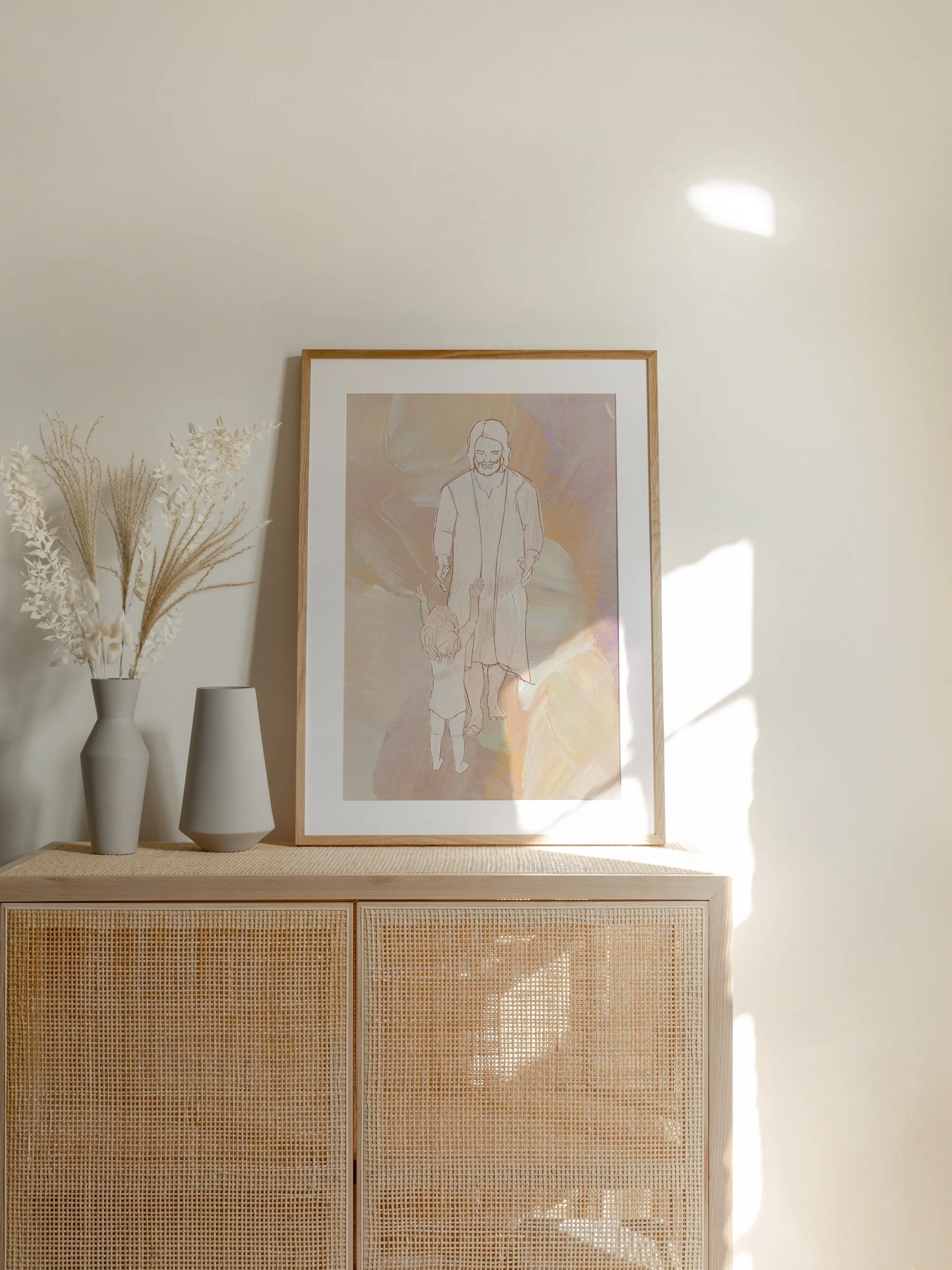 He Came Just for Me IV, framed in light wood with a white mat, standing on a rattan cabinet beside dried grasses and pale vases in sunlight.