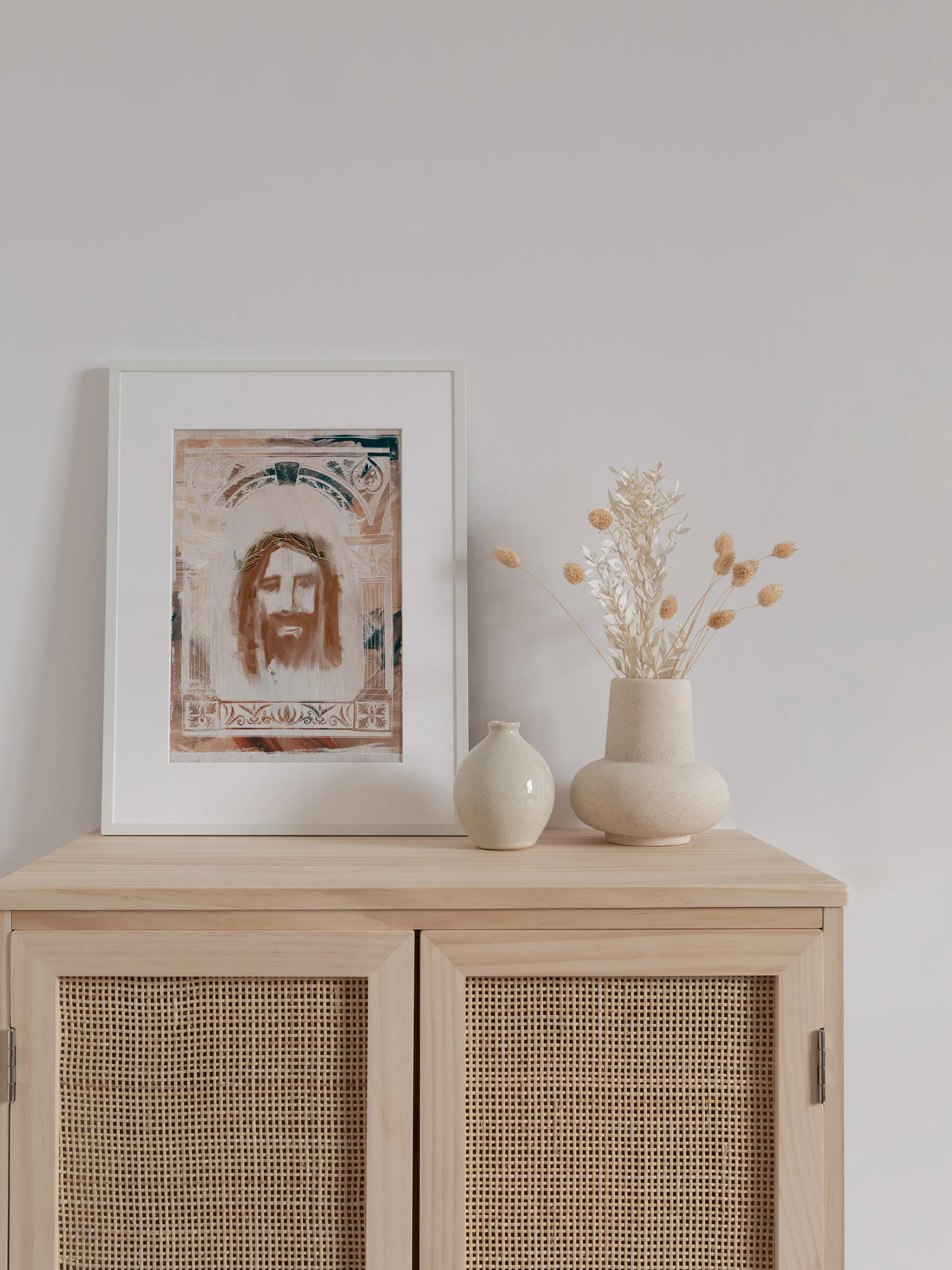 Jesus Crown of Thorns, Art Deco, framed in white with a white mat, standing on a light wood cabinet beside small vases and dried flowers.