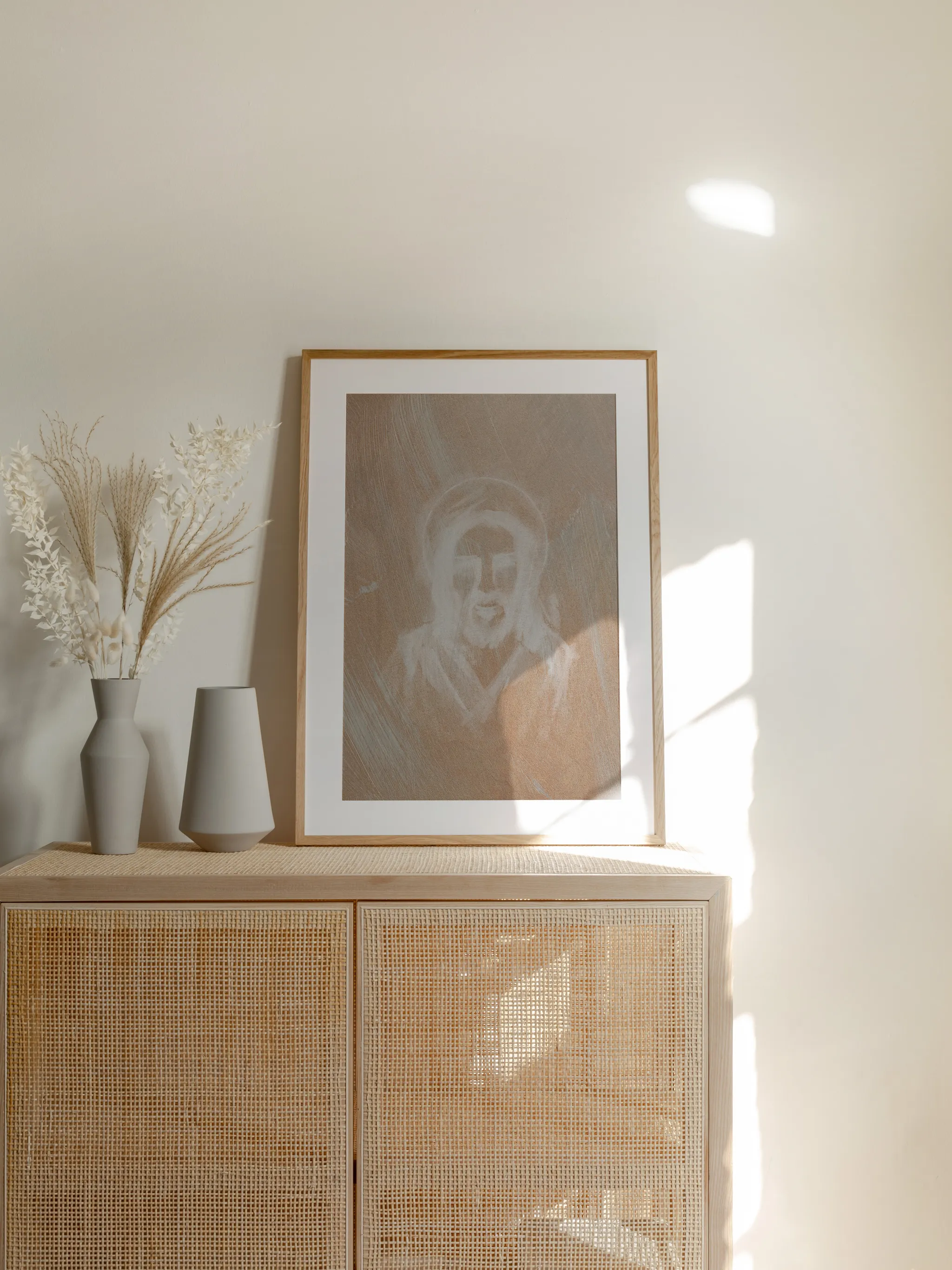 Jesus Portrait, Etched in White, framed in light wood with a white mat, standing on a rattan cabinet beside dried grasses and pale vases in sunlight.
