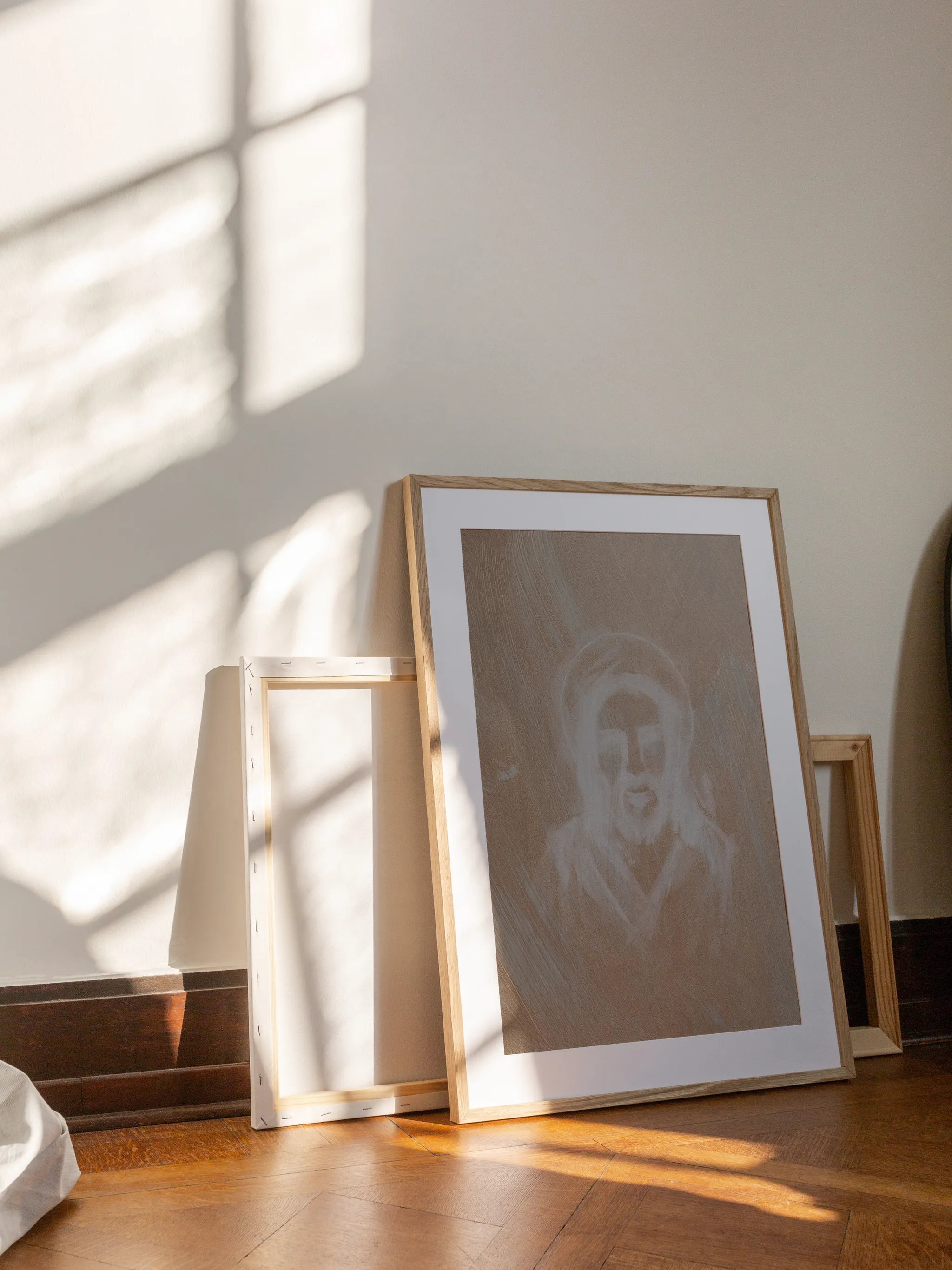 Jesus Portrait, Etched in White, framed in light wood with a white mat, leaning against a wall on a wooden floor in slanting sunlight, with empty frames behind it.