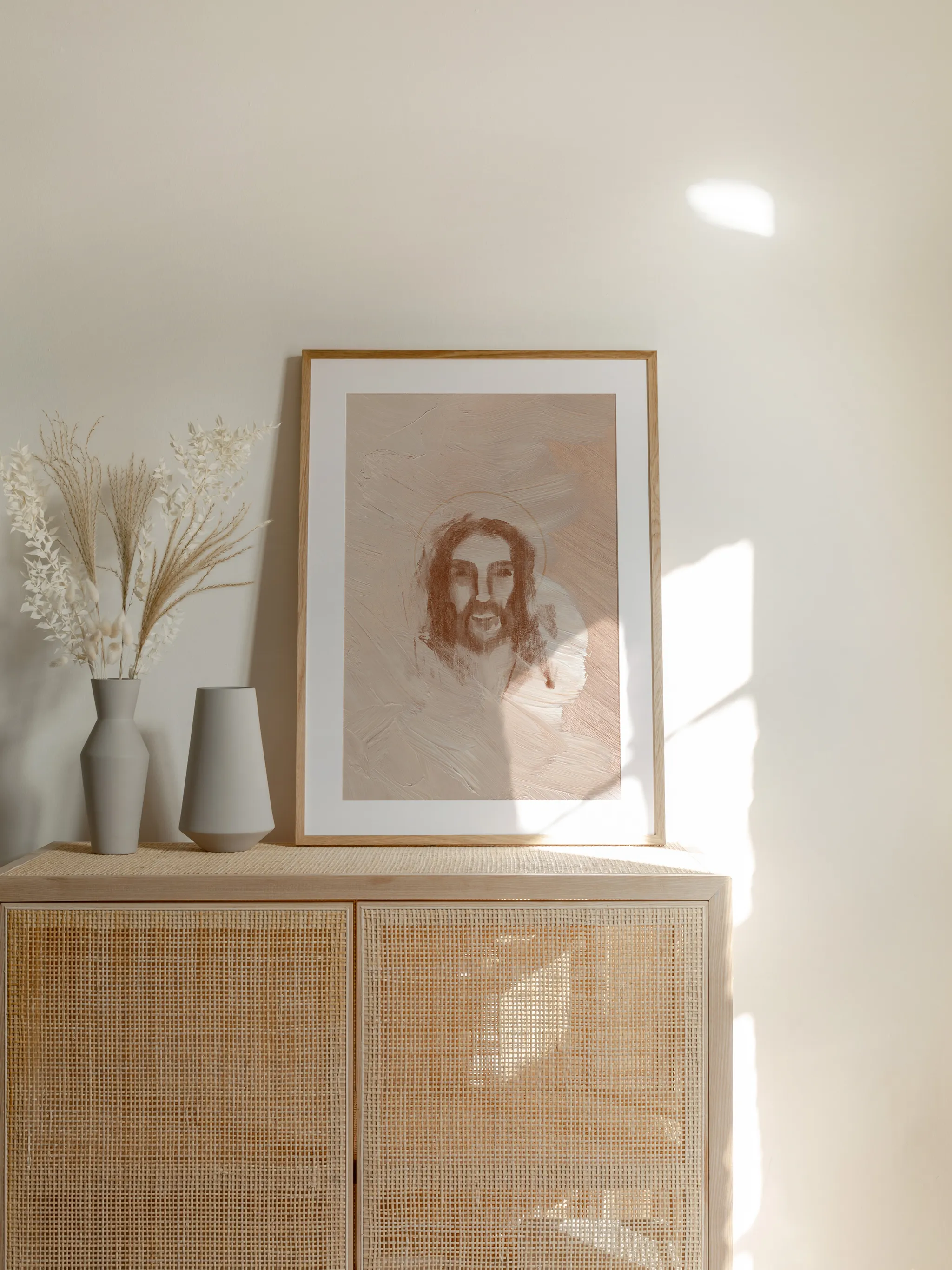 Jesus Portrait Watercolor II, framed in light wood with a white mat, standing on a rattan cabinet beside dried grasses and pale vases in sunlight.