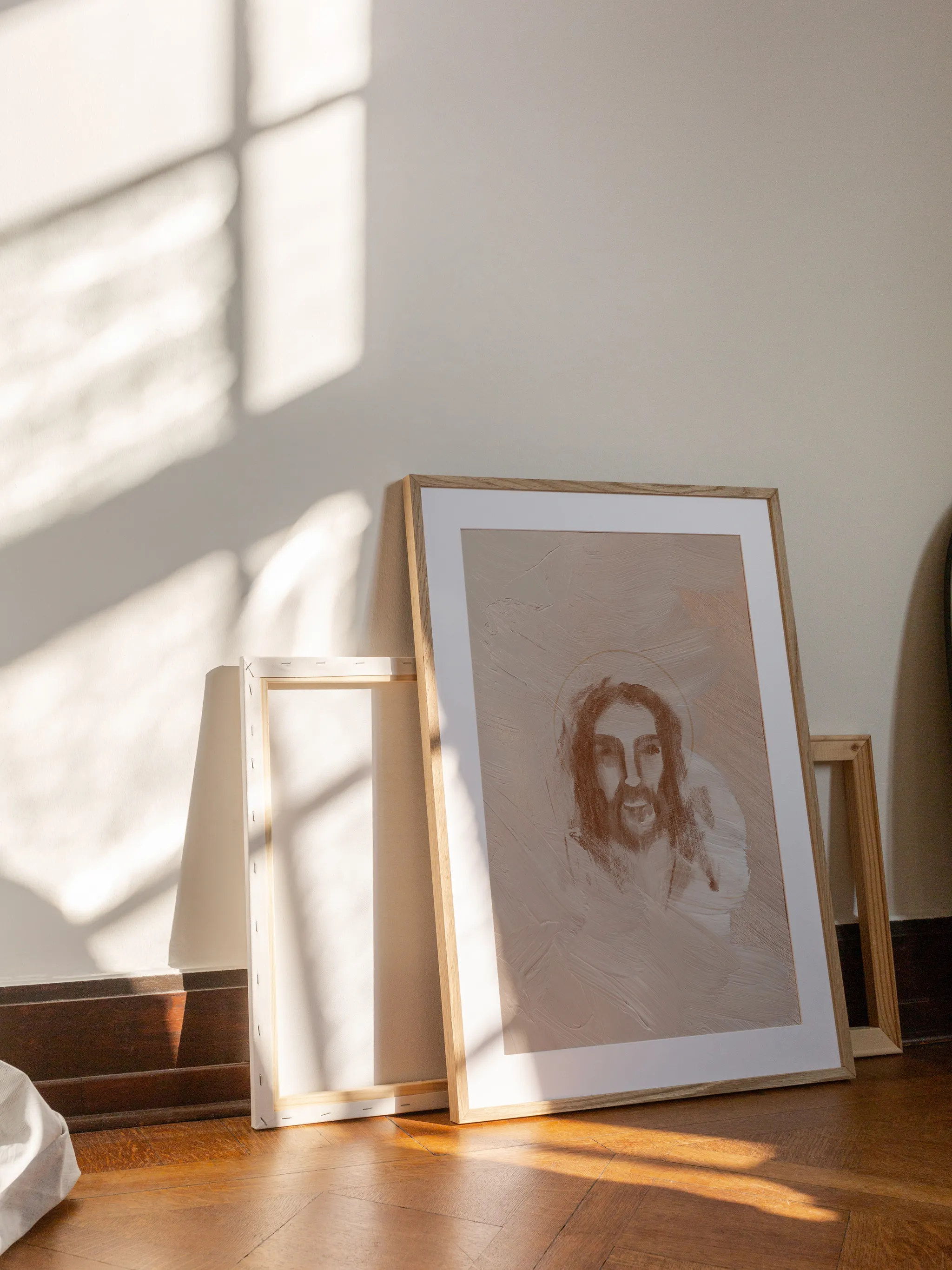 Jesus Portrait Watercolor II, framed in light wood with a white mat, leaning against a wall on a wooden floor in slanting sunlight, with empty frames behind it.