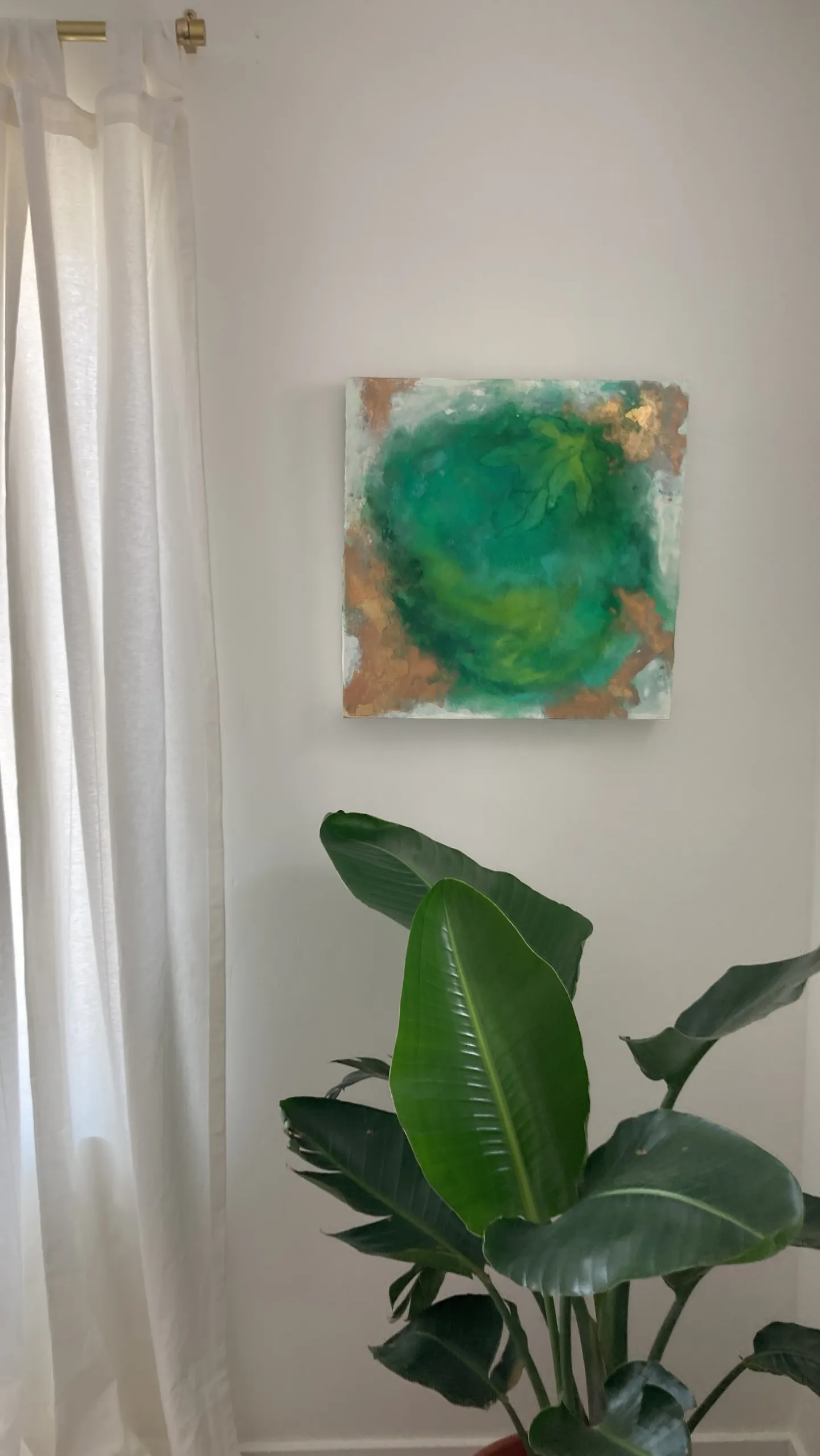 Mother Eve hanging on a white wall beside a sheer white curtain, above a potted bird of paradise.
