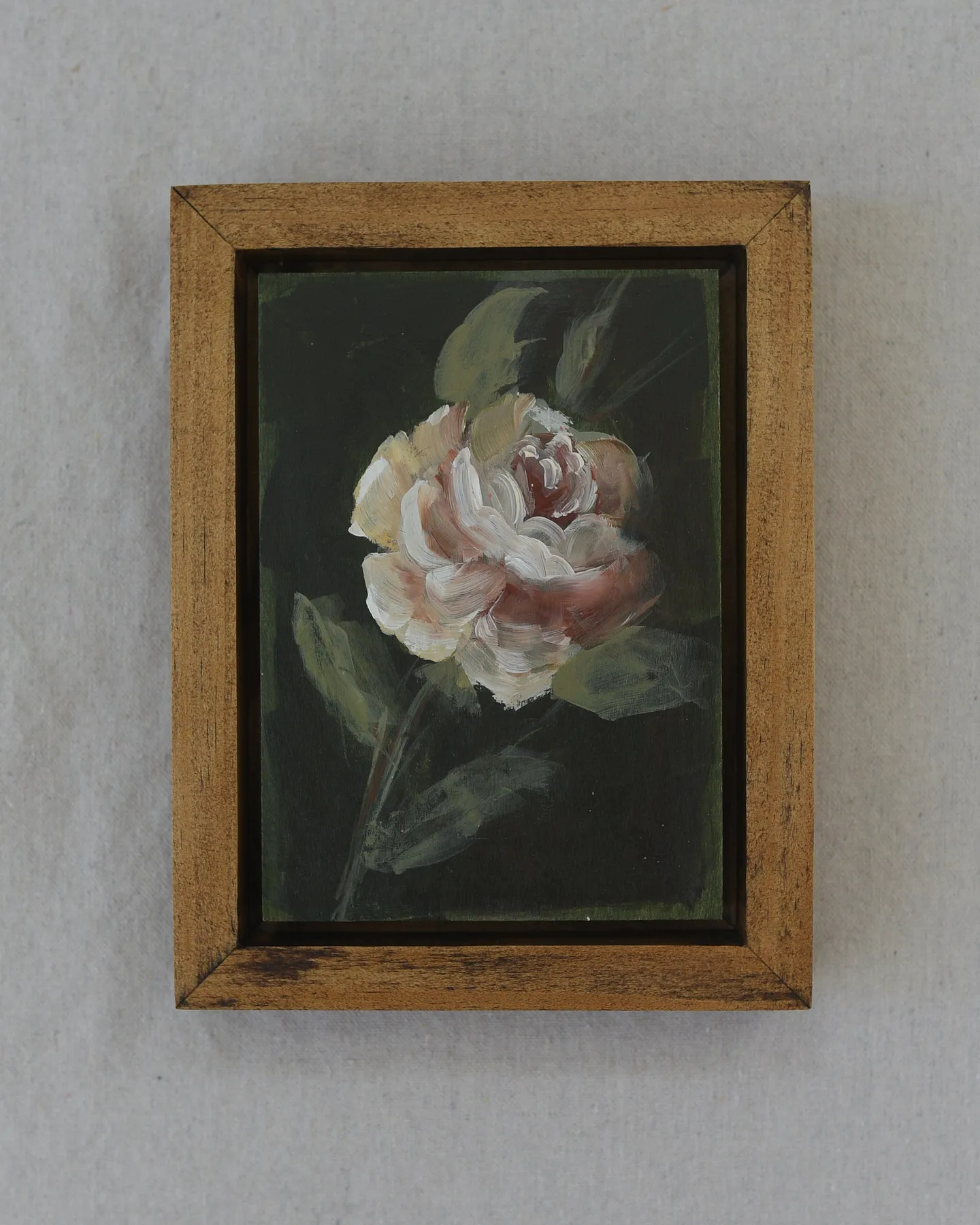 A delicate rose blooms against a dark olive green background, painted in acrylic on raw wood.
