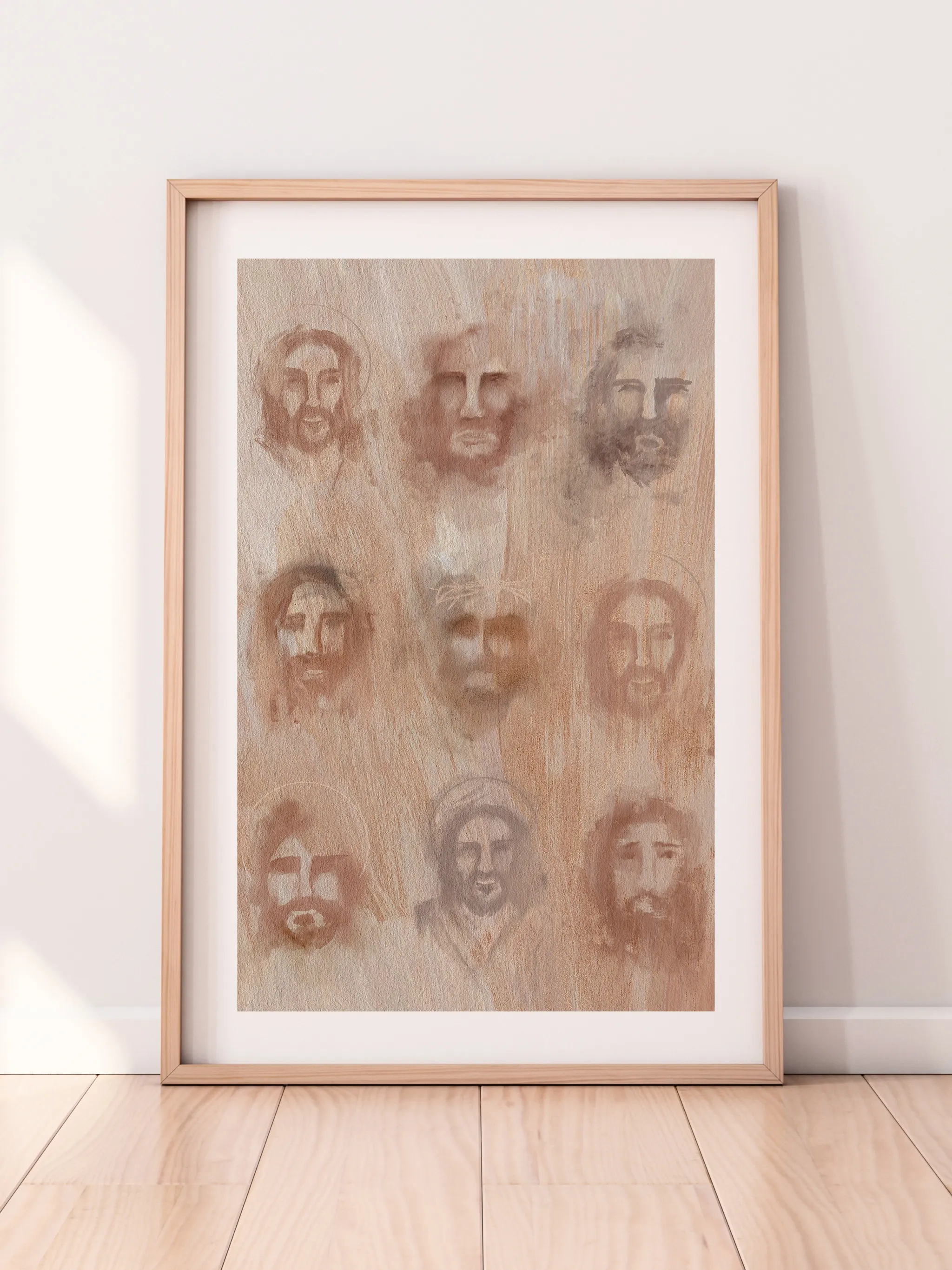 The Faces of Christ, framed in light wood with a white mat, leaning against a pale wall on a light wood floor.