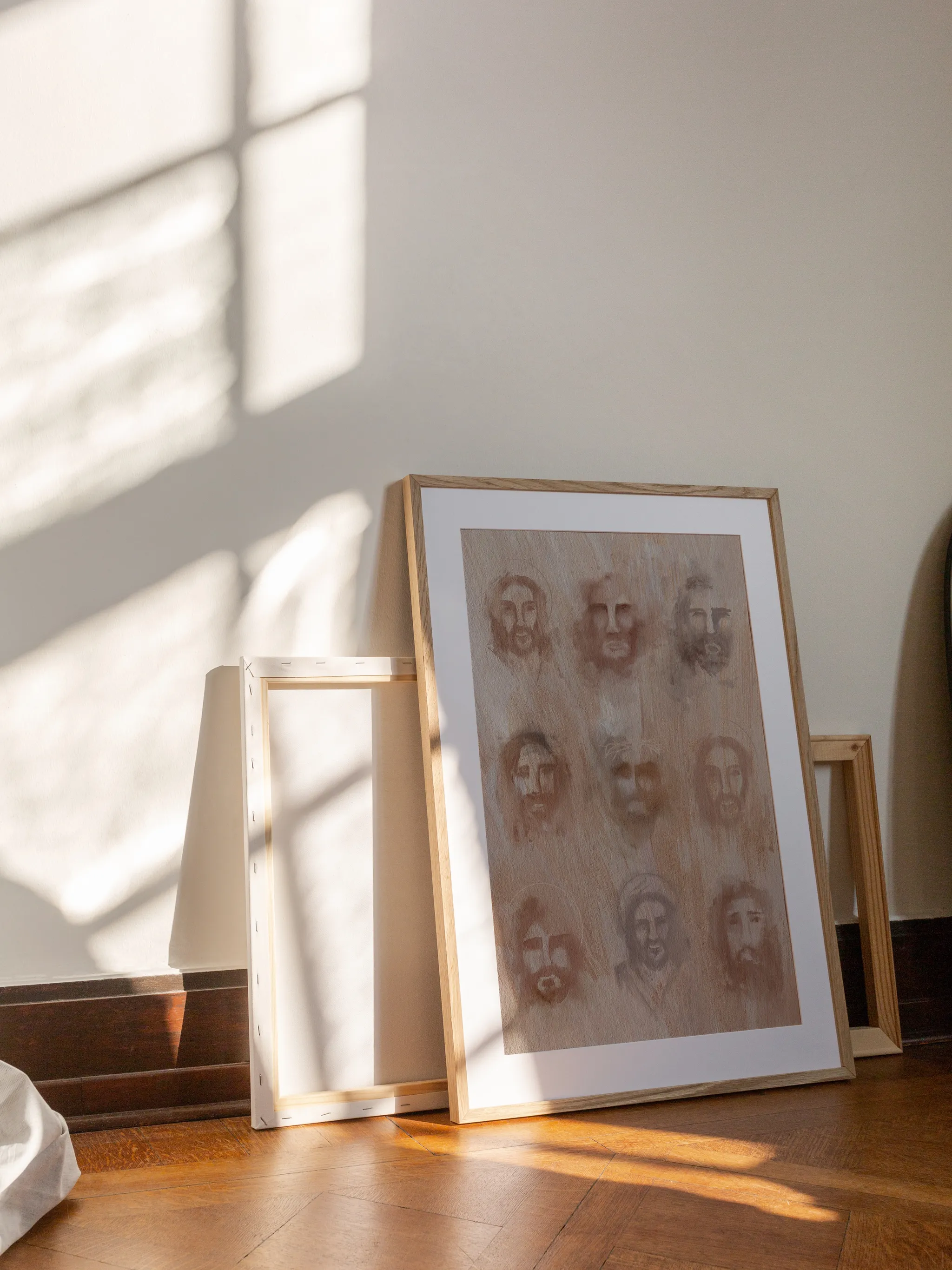The Faces of Christ, framed in light wood with a white mat, leaning against a wall on a wooden floor in slanting sunlight, with empty frames behind it.