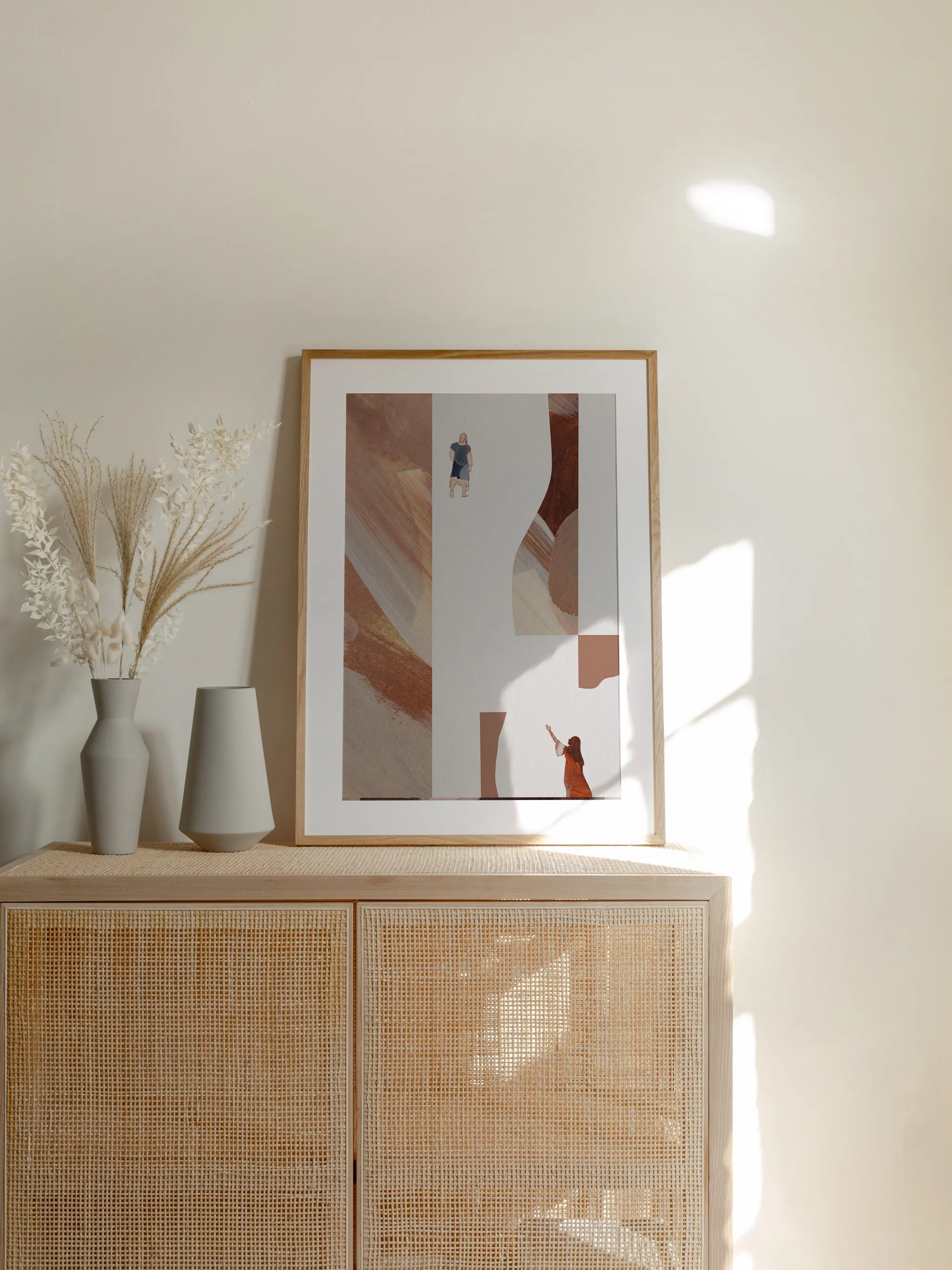 Woman with an Issue of Blood I, framed in light wood with a white mat, standing on a rattan cabinet beside dried grasses and pale vases in sunlight.