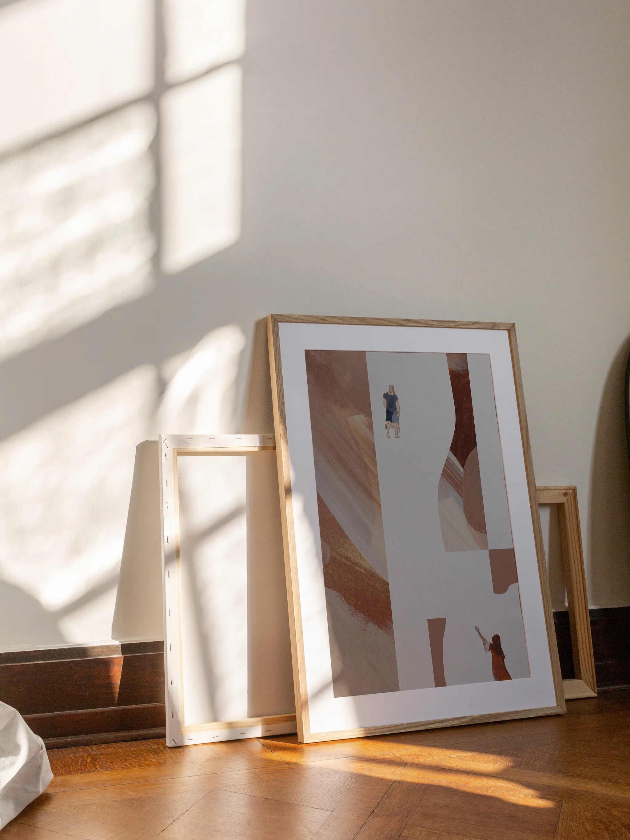 Woman with an Issue of Blood I, framed in light wood with a white mat, leaning against a wall on a wooden floor in slanting sunlight, with empty frames behind it.