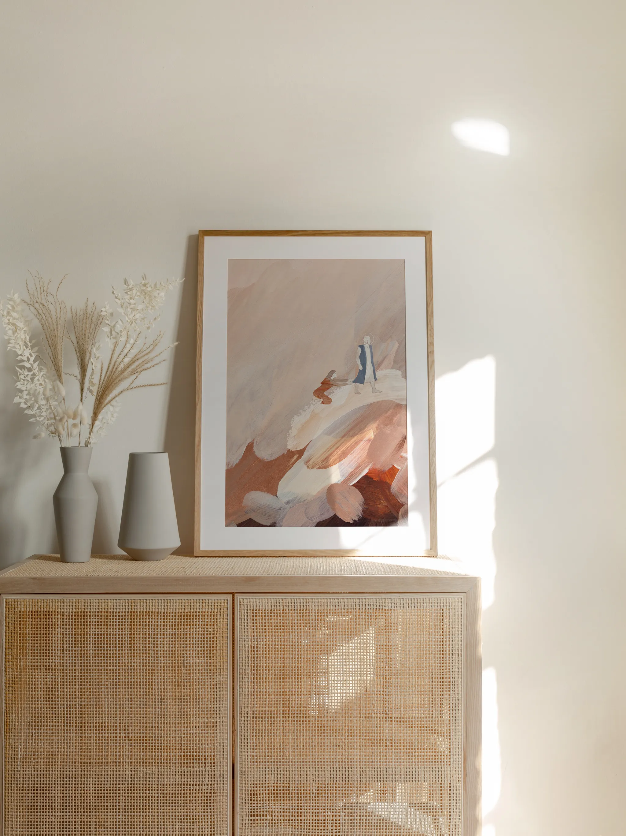 Woman with an Issue of Blood III, framed in light wood with a white mat, standing on a rattan cabinet beside dried grasses and pale vases in sunlight.