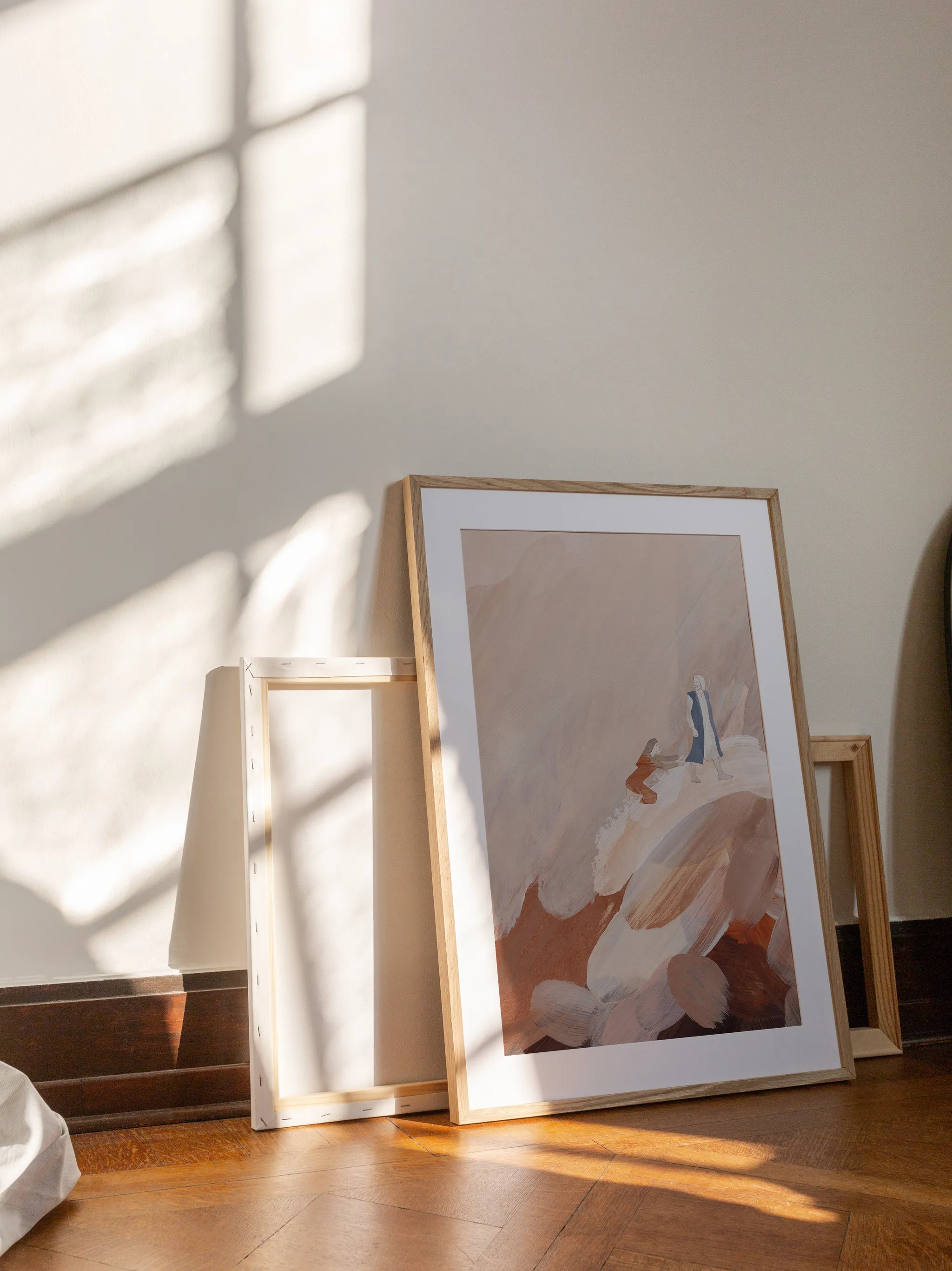 Woman with an Issue of Blood III, framed in light wood with a white mat, leaning against a wall on a wooden floor in slanting sunlight, with empty frames behind it.