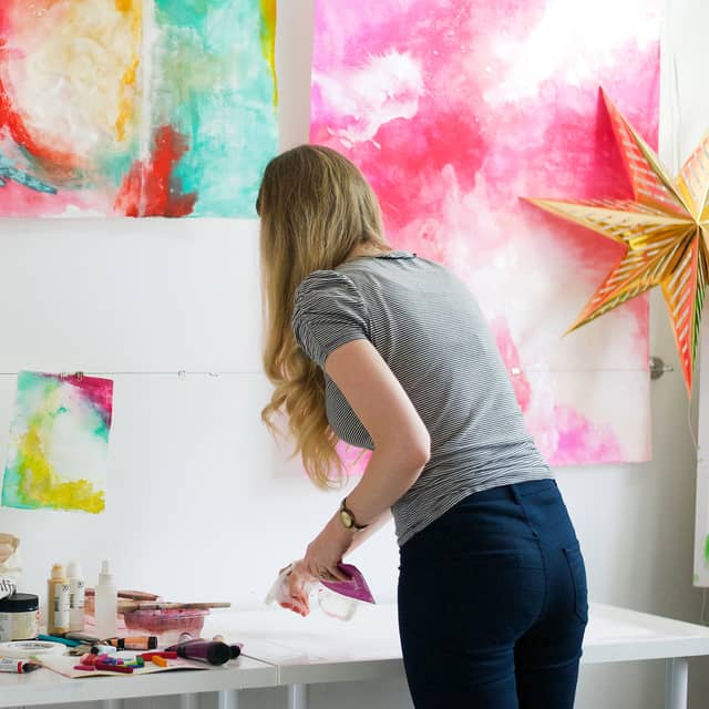 McKenna at her studio table with her back to us, working under a wall of bright pink and yellow abstracts.