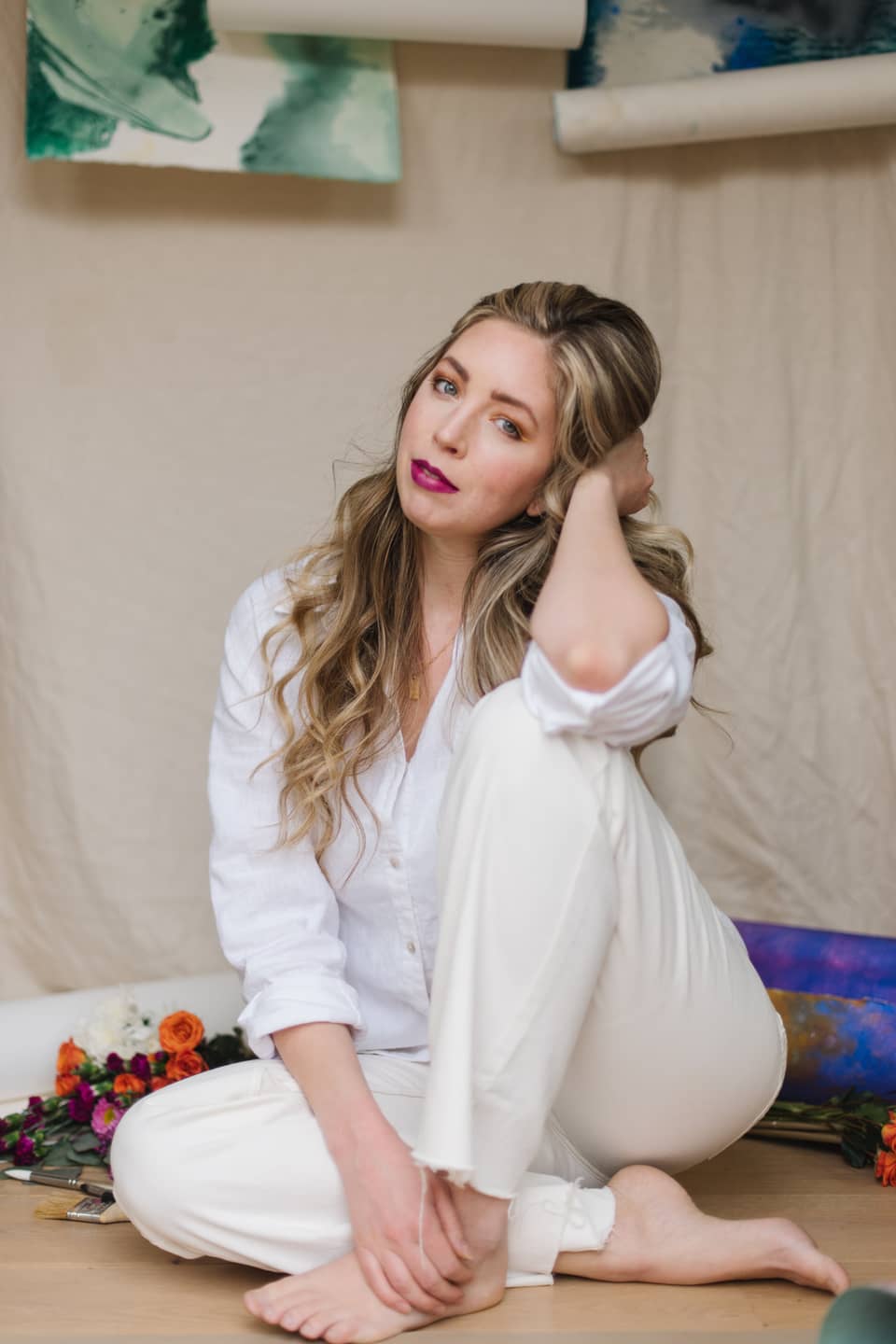McKenna sitting barefoot on the studio floor in white, with flowers and rolled canvases around her.