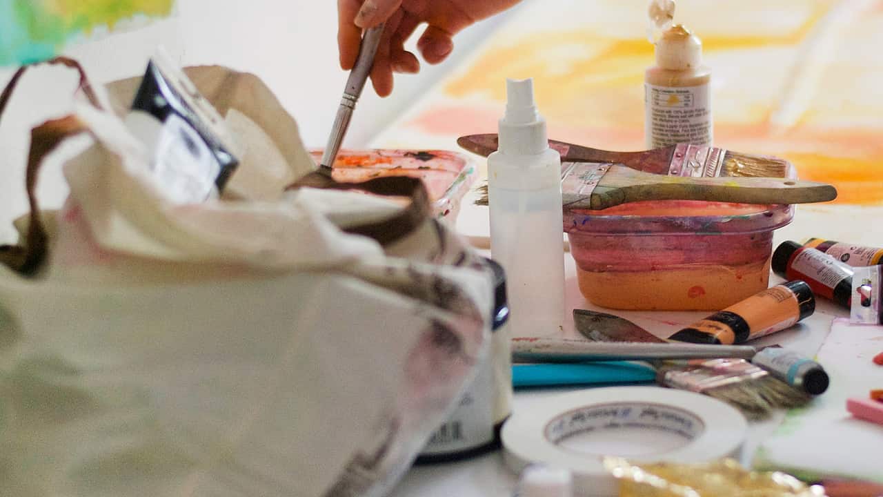 A hand mixing paint over a cluttered work table of brushes, jars and colour.