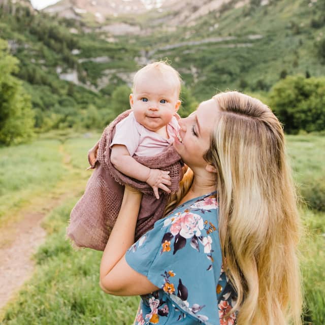 McKenna kissing her baby daughter, held up against a green mountain meadow.
