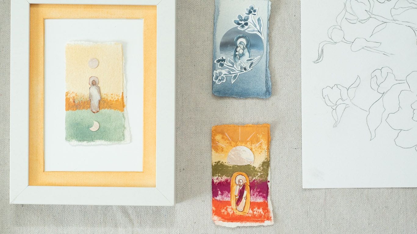 Small paintings laid out on linen: one framed in white with a marigold mat, a blue mother and child and a sunrise over bands of colour laid loose beside it, and a pencil drawing of flowers.
