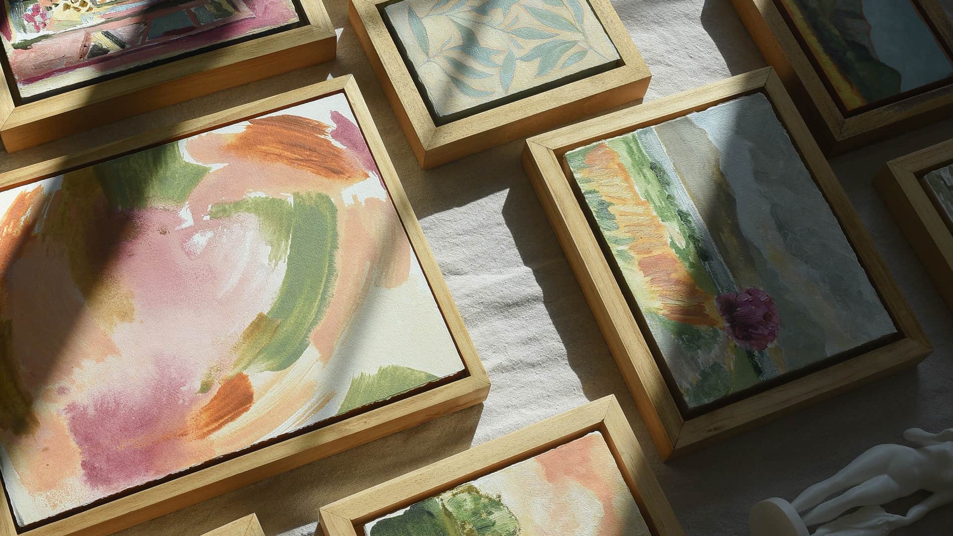 Fields & Flowers. Dozens of small paintings in raw wood float frames laid out together on linen, lit by a shaft of afternoon sun.