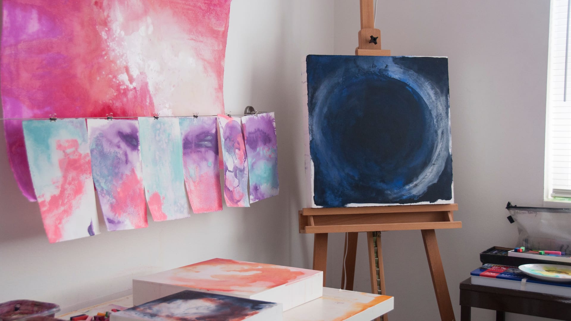 McKenna's studio with Overcome, the deep indigo circle, on a wooden easel, beside a row of pink and violet paper studies clipped to a wire above a table of painted canvases.