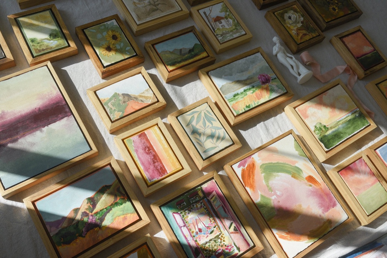 Dozens of small framed paintings, landscapes, florals and soft abstracts in raw wood float frames, laid out on linen in a shaft of sunlight.