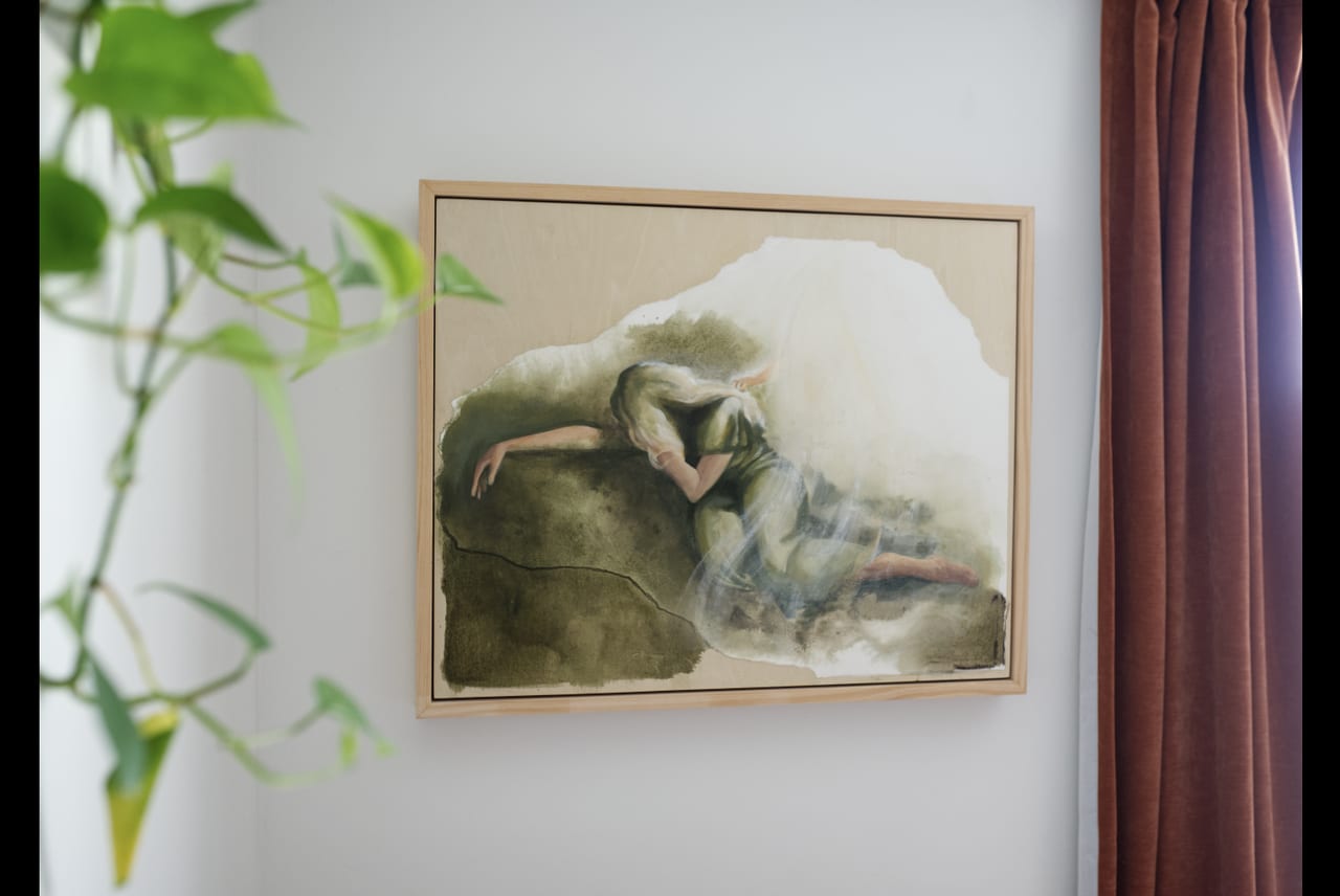 A Mother's Gethsemane framed in raw wood on a white wall, with a trailing green plant at one side and a deep terracotta curtain at the other.
