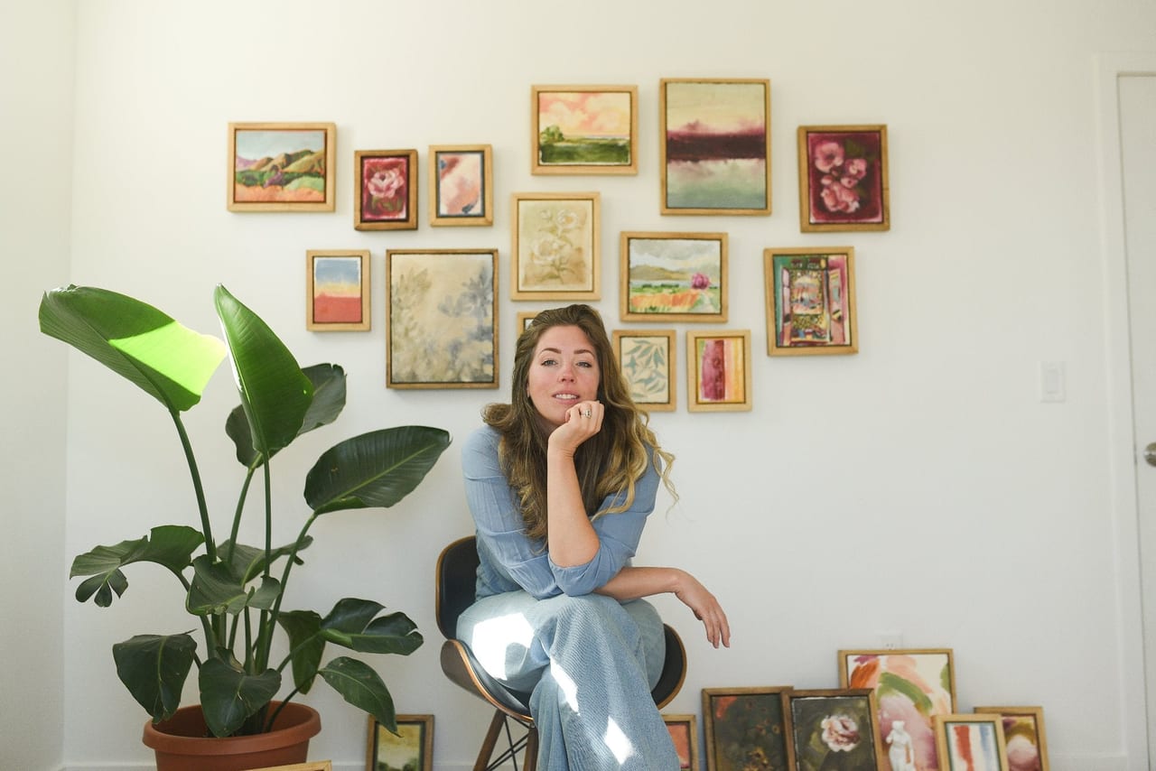 McKenna sitting in front of a wall hung with two dozen of her small framed paintings, a large-leaved plant beside her.