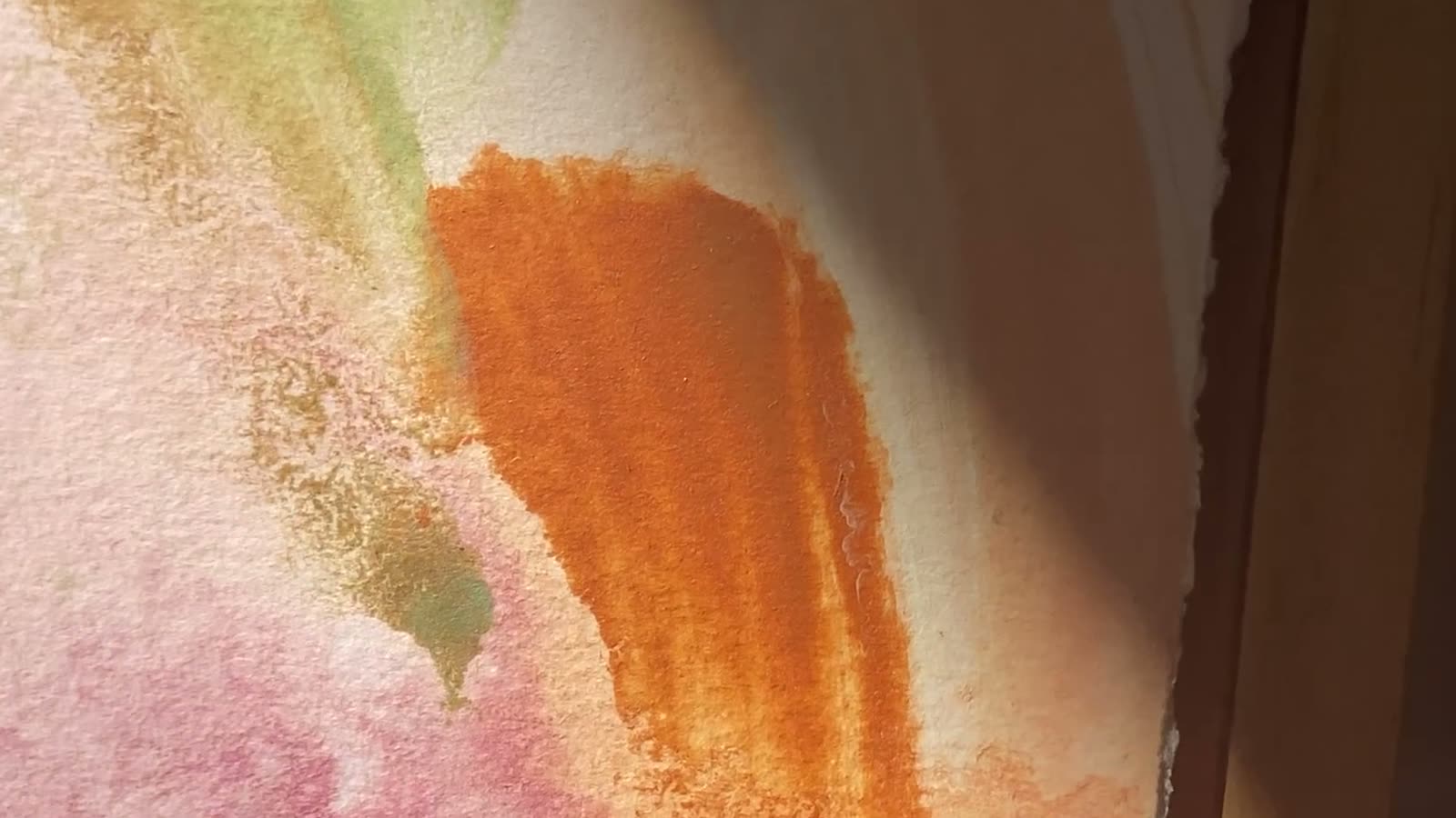 A close view of an abstract painting, pink, orange and green washes with a beam of sunlight falling across the paint.